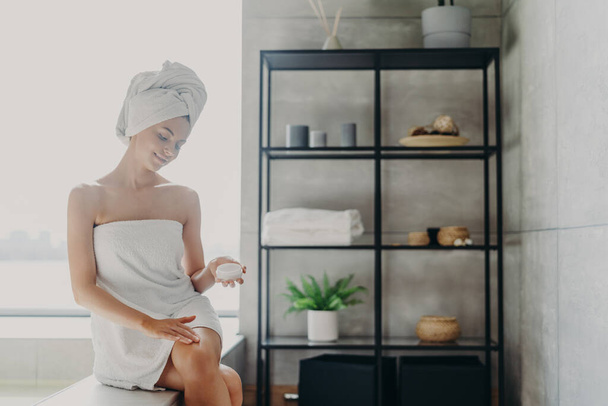 Photo of relaxed beautiful young woman applies body cream at home, uses cosmetic product for healthy skin, poses in bathroom, wearrs wrapped towel on head and over body. Beauty treatment concept - Photo, Image
