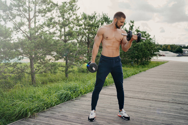 Sport and bodybuilding concept. Muscular bearded European man raises heavy barbells wears sport trousers and sneakers flexes muscles outdoors, does weightlifting exercises, leads active lifestyle - Photo, Image