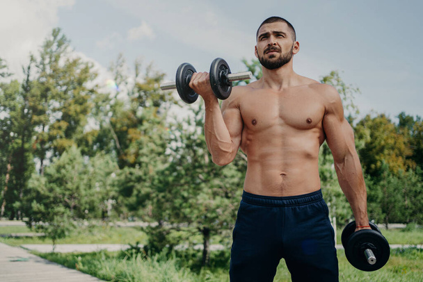 Horizontal shot of topless fitness unshaven European man has muscular body, raises barbells, wears shorts, demonstrates strong arms, has workout outdoor in park. Sportsman makes weightlifting - Foto, imagen