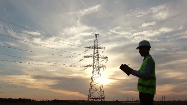 An engineer in special clothes makes notes on a tablet and turns to a high-voltage pole against the backdrop of a beautiful sunset. - Footage, Video