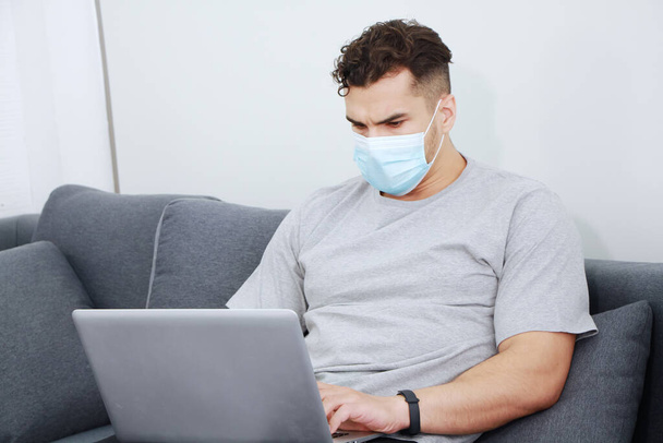 Νεαρός Καυκάσιος επιχειρηματίας φορώντας μάσκα εμποδίζει τον ιό covid-19 να χαλαρώσει και να συνεργαστεί με το laptop σε online μέσα κοινωνικής δικτύωσης στο σαλόνι στο σπίτι της οικογένειας. Η τεχνολογία και η εργασία αποτελούν έννοια του σπιτιού. - Φωτογραφία, εικόνα