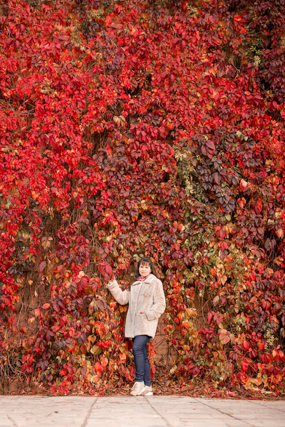 A girl in a beige coat stands next to a red wall of grapes in autumn. - Fotó, kép