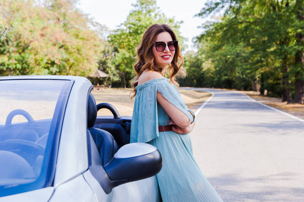 Happy pretty girl. Attractive woman leaning and posing at convertible car. Outdoor fashion portrait. Wearing elegant dress and sunglasses. Looking at camera. Enjoying beautiful summer day in nature after driving car. - Fotó, kép