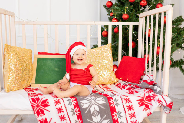 szczęśliwe małe dziecko sześć miesięcy w kapeluszu Mikołaja siedzi w łóżeczku w domu przy choince z tablicy tekstowej, nowy rok i koncepcja święta - Zdjęcie, obraz