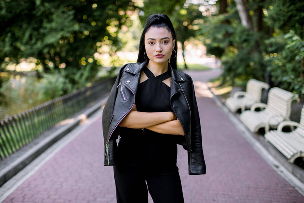 People, race, ethnicity and portrait concept. Horizontal photo of happy pretty mixed raced Asian lady in black overalls and leather jacket, posing to camera with crossed arms, walking in summer park - Zdjęcie, obraz
