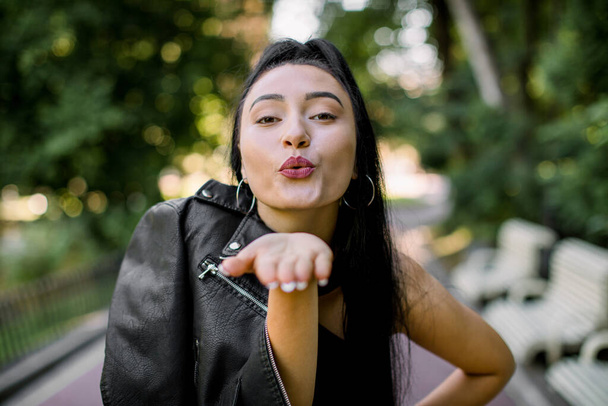 Amazing smiling Asian mixed raced model woman in black leather jacket, posing with different emotions at beautiful public park, showing air kiss to camera. Pretty woman enjoying a day at park - Foto, Bild