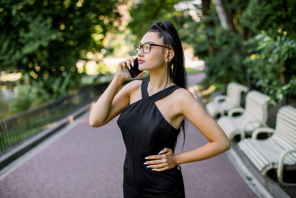 Young Chinese businesswoman with long black ponytail hair, looking worried and concentrated, having call with smart cell phone in city park outdoor. New technology trends concept. People portraits - Zdjęcie, obraz