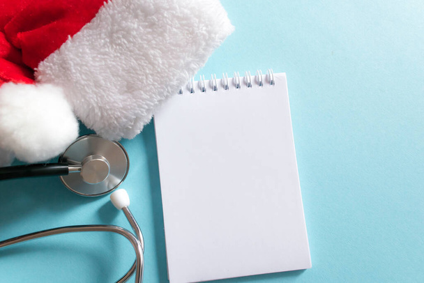 Medical Christmas and New year composition with stethoscope, notepad and Santa hat on blue background. Health care and medicine concept for card, calendar, cover. Copy space for text, flat lay. - Fotó, kép