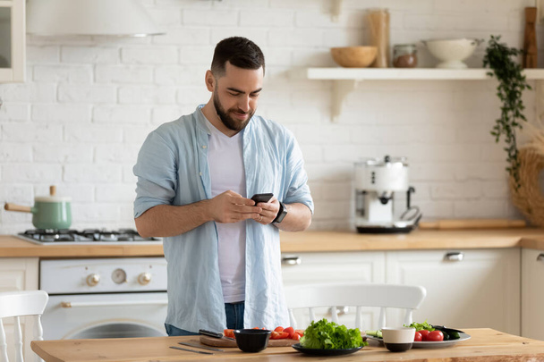 Young man using smartphone, cooking salad in kitchen, searching recipe - Foto, Imagem