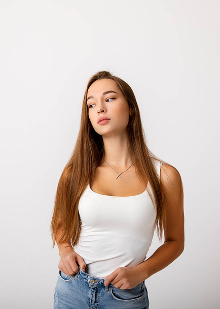 A beautiful young slender long haired brunette girl holds her jeans and looks thoughtfully to the side. The concept of healthy food, diet, lifestyle. Studio photo on white background - Foto, Bild