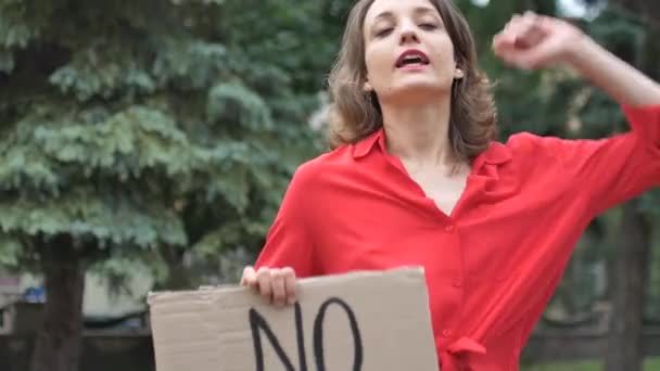 Mujer joven protestando enojada con camisa roja sostiene cartel de protesta con pancarta con el lema No hay racismo para la manifestación pública en el fondo de los árboles. - Metraje, vídeo