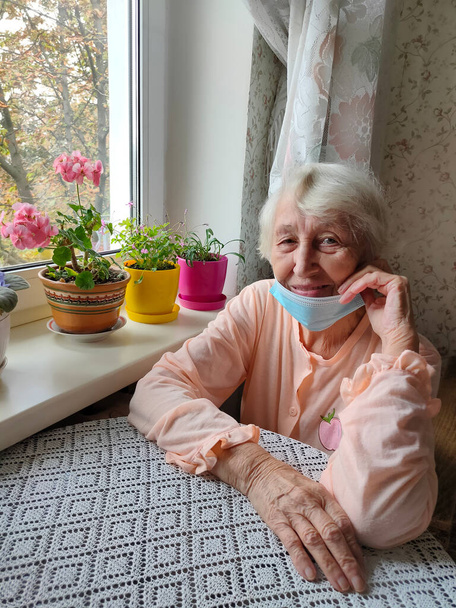 Druga fala. Covid-19, koncepcja zdrowia, bezpieczeństwa i pandemii - starsza samotna kobieta nosząca ochronną maskę medyczną siedząca w pobliżu okna w domu dla ochrony przed wirusem - Zdjęcie, obraz