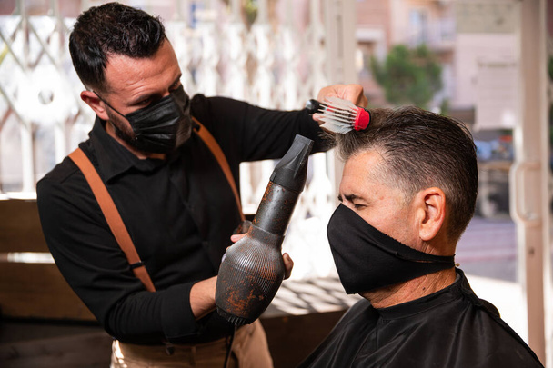 El peluquero blanco caucásico peina y seca el cabello de otro hombre en una barbería durante la pandemia del coronavirus. van estilo hipster retro y ambos usan máscaras negras. - Foto, imagen