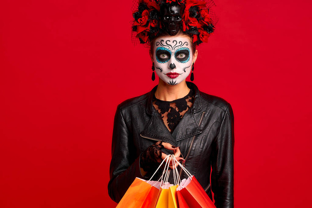 Girl with sugar skull makeup with a wreath of flowers on her head and skull, wearth lace gloves and leather jacket, hold shopper bags party preparation isolated on red background. - Foto, Bild