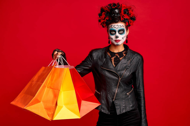 Dziewczyna z cukrem makijaż czaszki z wieńcem kwiatów na głowie i czaszce, nosić rękawiczki koronkowe i skórzaną kurtkę, trzymać torby shopper party przygotowanie izolowane na czerwonym tle. - Zdjęcie, obraz