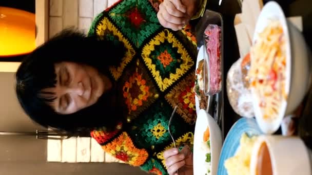 Голодная взрослая женщина кавказской национальности в яркой одежде, жадно кушающая много вкусной и разнообразной пищи, сидящая за столом в столовой или ресторане. Вертикальное видео. Средний план. - Кадры, видео