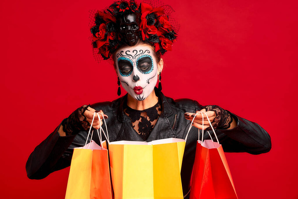 Girl with sugar skull makeup with a wreath of flowers on her head and skull, wearth lace gloves and leather jacket, hold shopper bags party preparation isolated on red background. - Photo, Image
