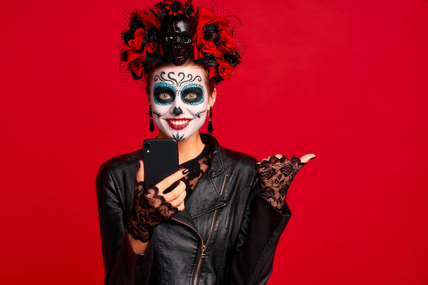 Girl with sugar skull makeup with a wreath of flowers on her head and skull, wearth lace gloves and leather jacket, shocked with news from her phone isolated on red background. - Photo, Image