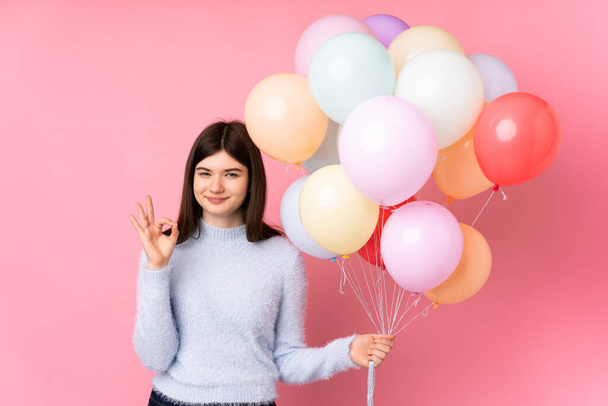 Young Ukrainian teenager girl holding lots of balloons over isolated pink background showing an ok sign with fingers - Photo, Image