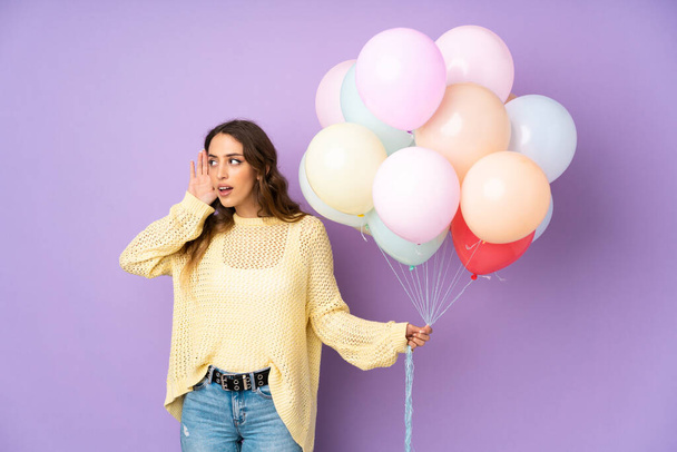 Νεαρή γυναίκα πιάνει πολλά μπαλόνια πάνω απομονωμένη σε μωβ φόντο ακούγοντας κάτι - Φωτογραφία, εικόνα