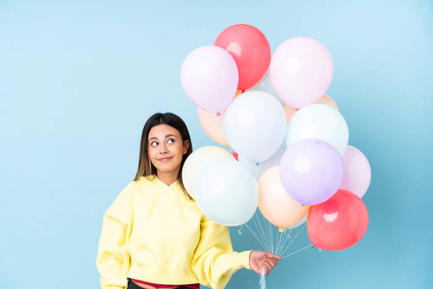 Frau hält Luftballons in einer Party über isoliertem blauem Hintergrund stehend und zur Seite blickend - Foto, Bild