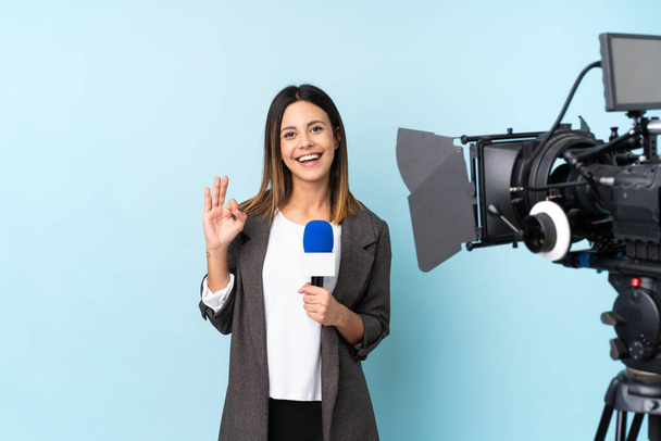 Δημοσιογράφος γυναίκα κρατώντας ένα μικρόφωνο και αναφέροντας ειδήσεις πάνω από απομονωμένο μπλε φόντο δείχνει μια πινακίδα ok με τα δάχτυλα - Φωτογραφία, εικόνα