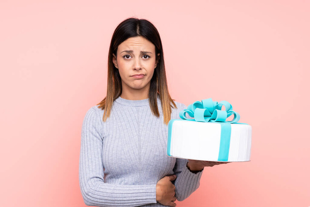 Woman holding a big cake over isolated pink background with sad expression - Fotografie, Obrázek
