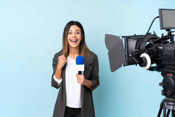 Reportera mujer sosteniendo un micrófono y reportando noticias sobre aislado fondo azul celebrando una victoria - Foto, imagen