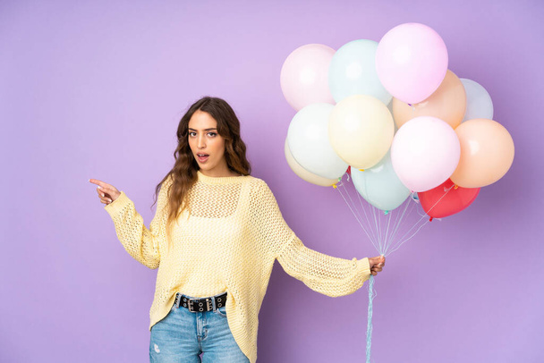 Young woman catching many balloons over isolated on purple background surprised and pointing finger to the side - Photo, Image