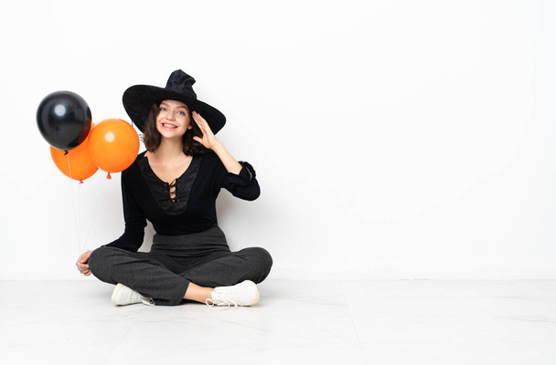 Young witch holding black and orange air balloons sitting on the floor with surprise expression - Photo, Image