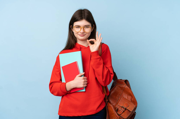 Jeune adolescente ukrainienne étudiante tenant une salade sur fond bleu isolé montrant un signe ok avec les doigts - Photo, image