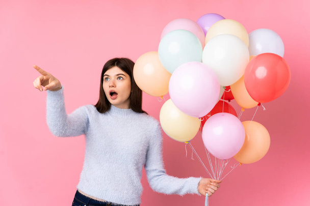 jong Oekraïense tiener meisje houden veel ballonnen over geïsoleerde roze achtergrond wijzend vinger naar de zijkant - Foto, afbeelding