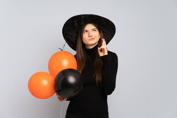 Młoda czarownica trzyma czarne i pomarańczowe balony z palcami skrzyżowanymi i życzy jak najlepiej - Zdjęcie, obraz
