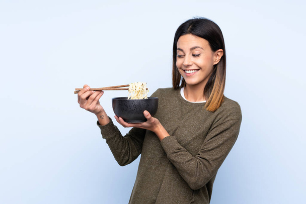 vrouw over geïsoleerde blauwe achtergrond met gelukkige uitdrukking terwijl het houden van een kom noedels met eetstokjes - Foto, afbeelding