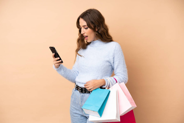 Молодая женщина на изолированном фоне держит сумки с покупками и пишет сообщение с мобильного телефона другу - Фото, изображение