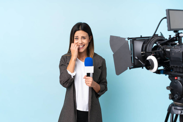 Reportera mujer sosteniendo un micrófono y reportando noticias sobre aislado fondo azul nervioso y asustado - Foto, imagen