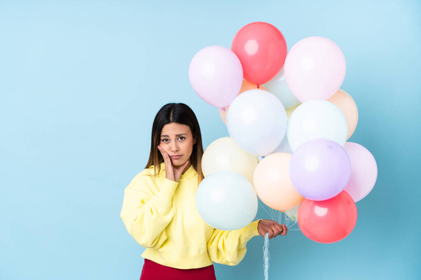 Frau hält Luftballons in einer Party über isoliertem blauem Hintergrund unglücklich und frustriert - Foto, Bild