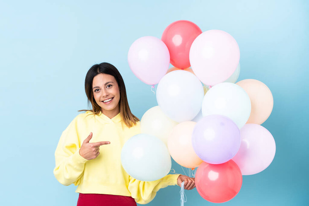 Woman holding balloons in a party over isolated blue background pointing finger to the side - Foto, afbeelding