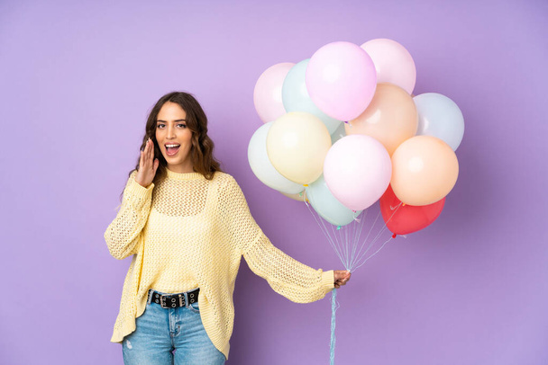Young woman catching many balloons over isolated on purple background shouting with mouth wide open - 写真・画像