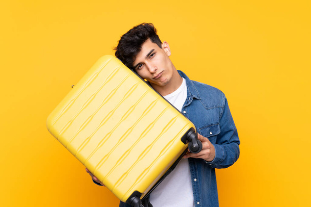 Jeune Argentin sur fond jaune isolé en vacances avec valise de voyage et malheureux - Photo, image