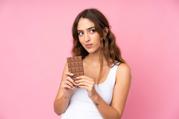 Young woman over isolated pink background taking a chocolate tablet and having doubts - Фото, изображение