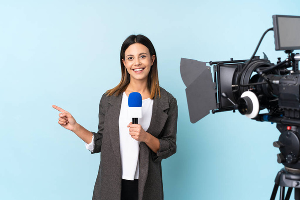 Женщина-репортер держит микрофон и сообщает новости на изолированном синем фоне, указывая пальцем в сторону - Фото, изображение