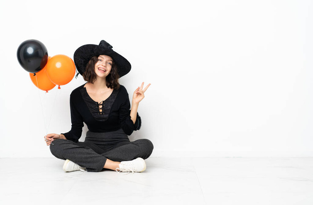 Молодая ведьма с черными и оранжевыми воздушными шарами сидит на полу улыбаясь и показывая знак победы - Фото, изображение
