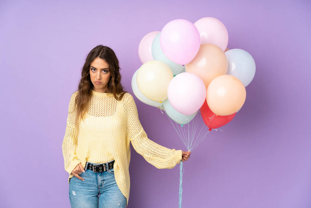 Young woman catching many balloons over isolated on purple background sad - Photo, Image
