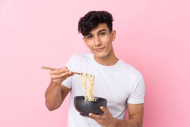 Young Argentinian man over isolated white background holding a bowl of noodles with chopsticks and offering it - Photo, Image