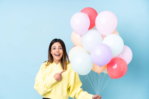 Woman holding balloons in a party over isolated blue background pointing finger to the side - Φωτογραφία, εικόνα