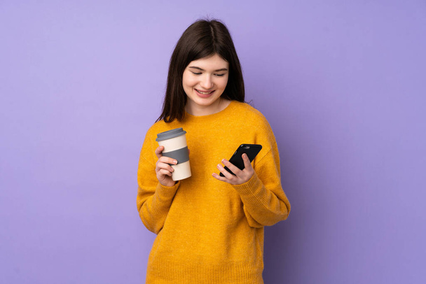 Νεαρή Ουκρανή έφηβη κοπέλα πάνω από απομονωμένο μωβ φόντο κρατώντας καφέ για να πάρει μακριά και ένα κινητό - Φωτογραφία, εικόνα