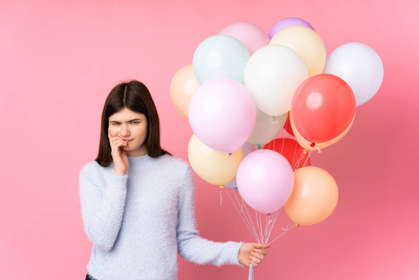 jong Oekraïense tiener meisje houden veel ballonnen over geïsoleerde roze achtergrond nerveus en bang - Foto, afbeelding