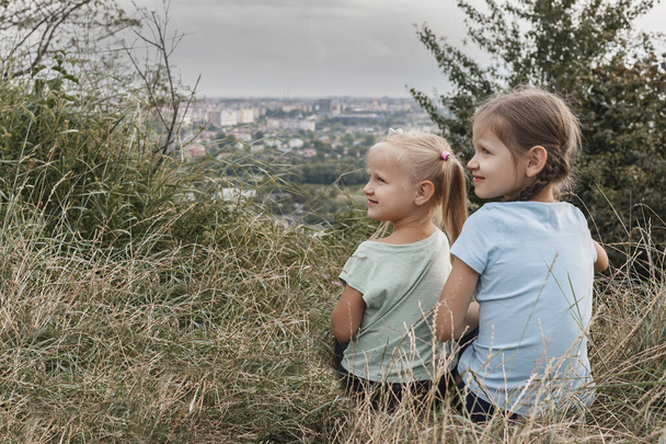 Zwei kleine Mädchen sitzen nebeneinander im Gras und schauen weg. Zwei Schwestern auf einem Hügel über der Stadt - Foto, Bild