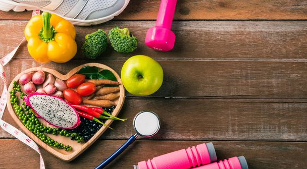 Вид зверху на свіжі органічні фрукти та овочі в серцевій пластині, взуття, спортивне обладнання та стетоскоп лікаря, студійний знімок на дерев'яному тренажерному столі, концепція здорової дієти вегетаріанської їжі, Всесвітній день їжі
 - Фото, зображення